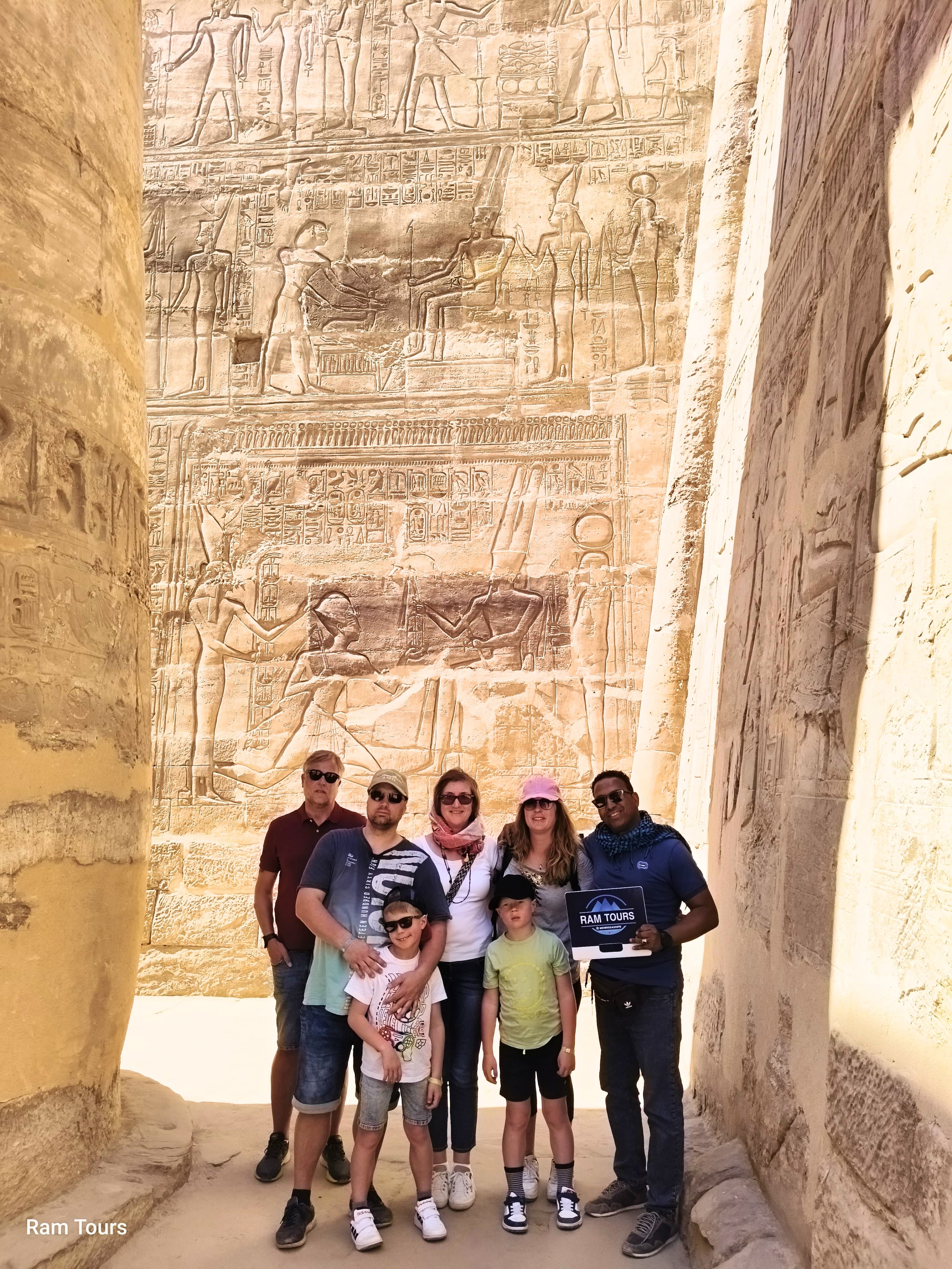 A group of tourists posing in front of a massive wall of ancient Egyptian hieroglyphics.