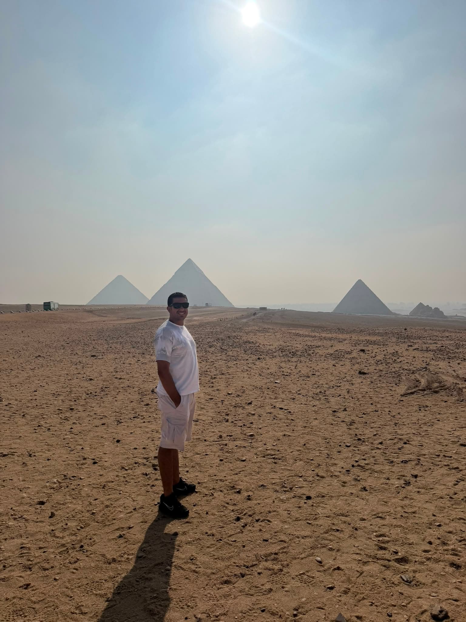 Man in white clothing stands in a desert with the three Pyramids of Giza behind.