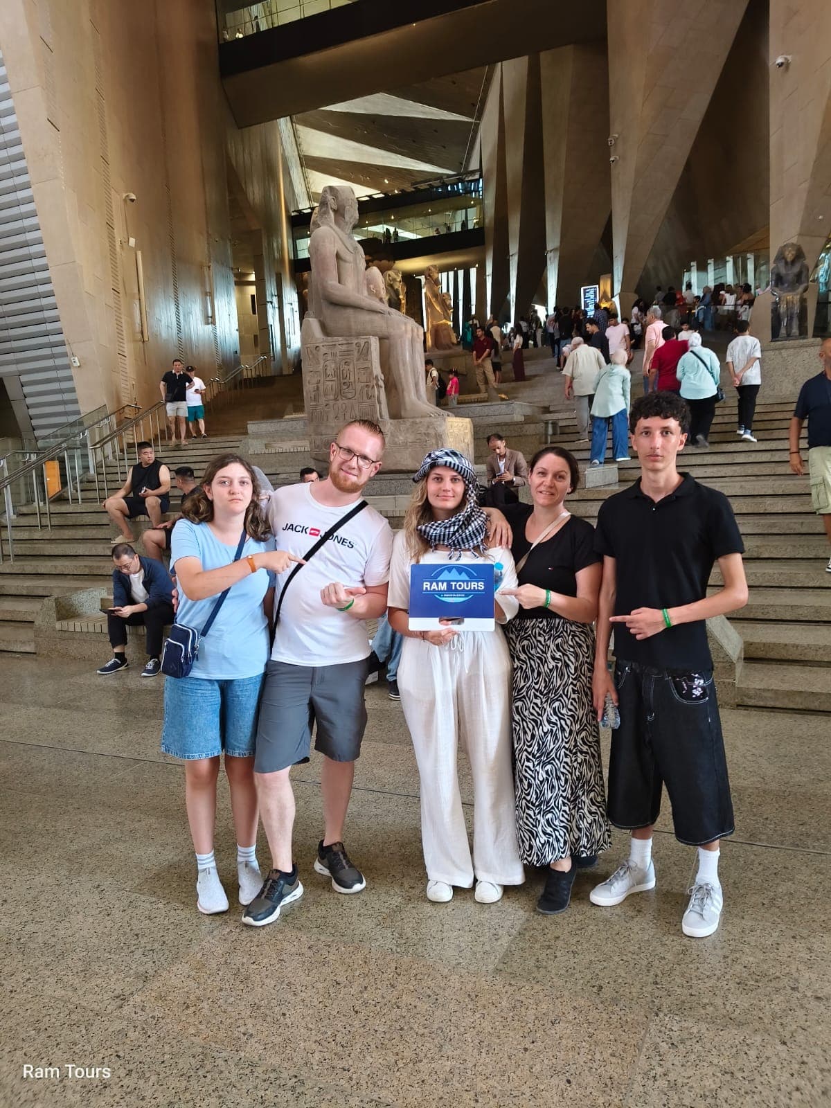 Tourists holding a Ram Tours sign pose before a colossal Egyptian statue in a museum.