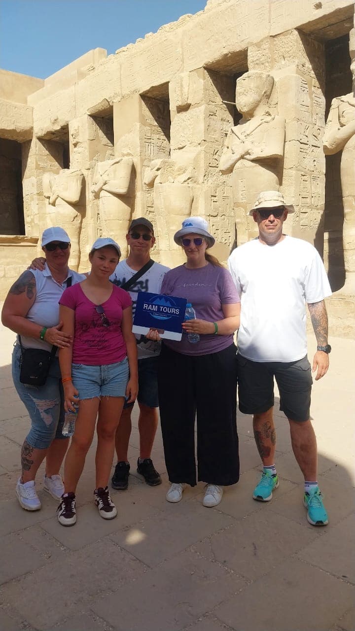 Five tourists pose with a tour sign in front of ancient Egyptian statues and hieroglyphics.