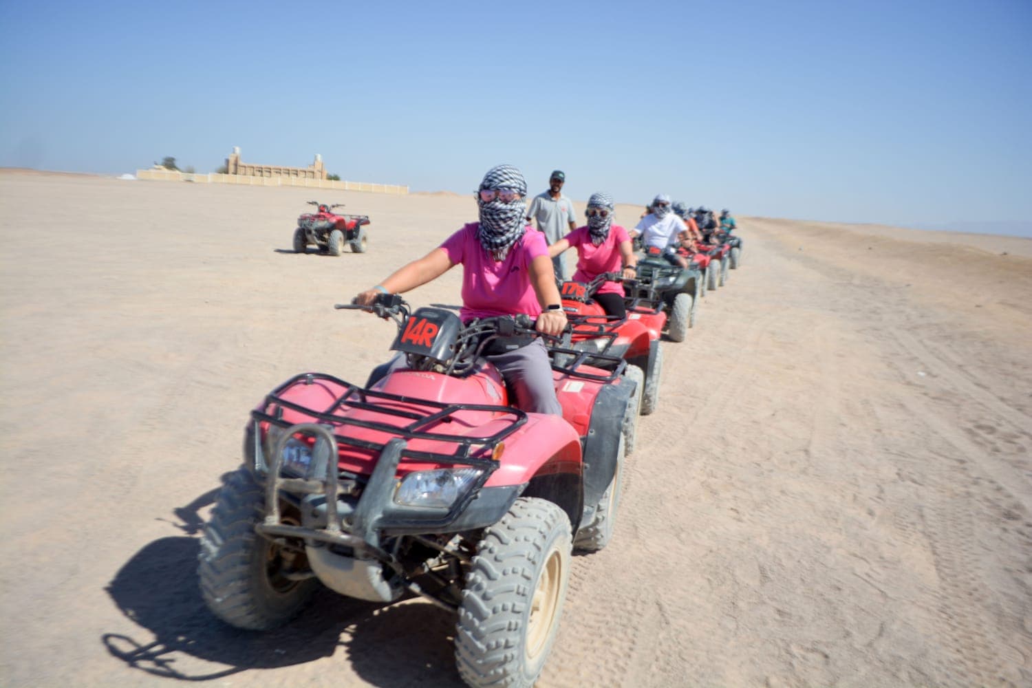 Tourists in headscarves ride red quad bikes in a line across a vast sandy desert.