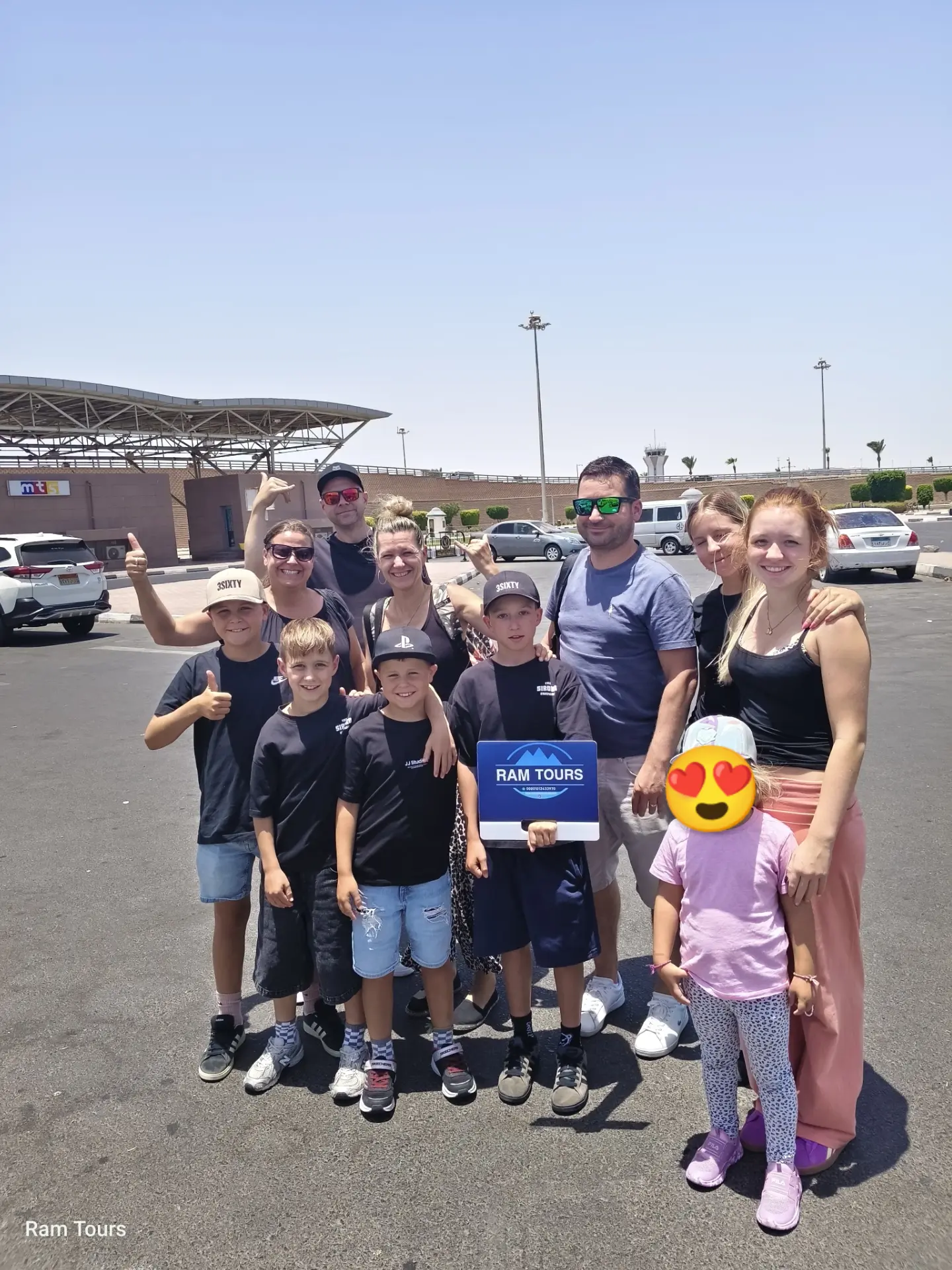 Smiling group of tourists posing with a RAM TOURS sign in a sunny parking lot.