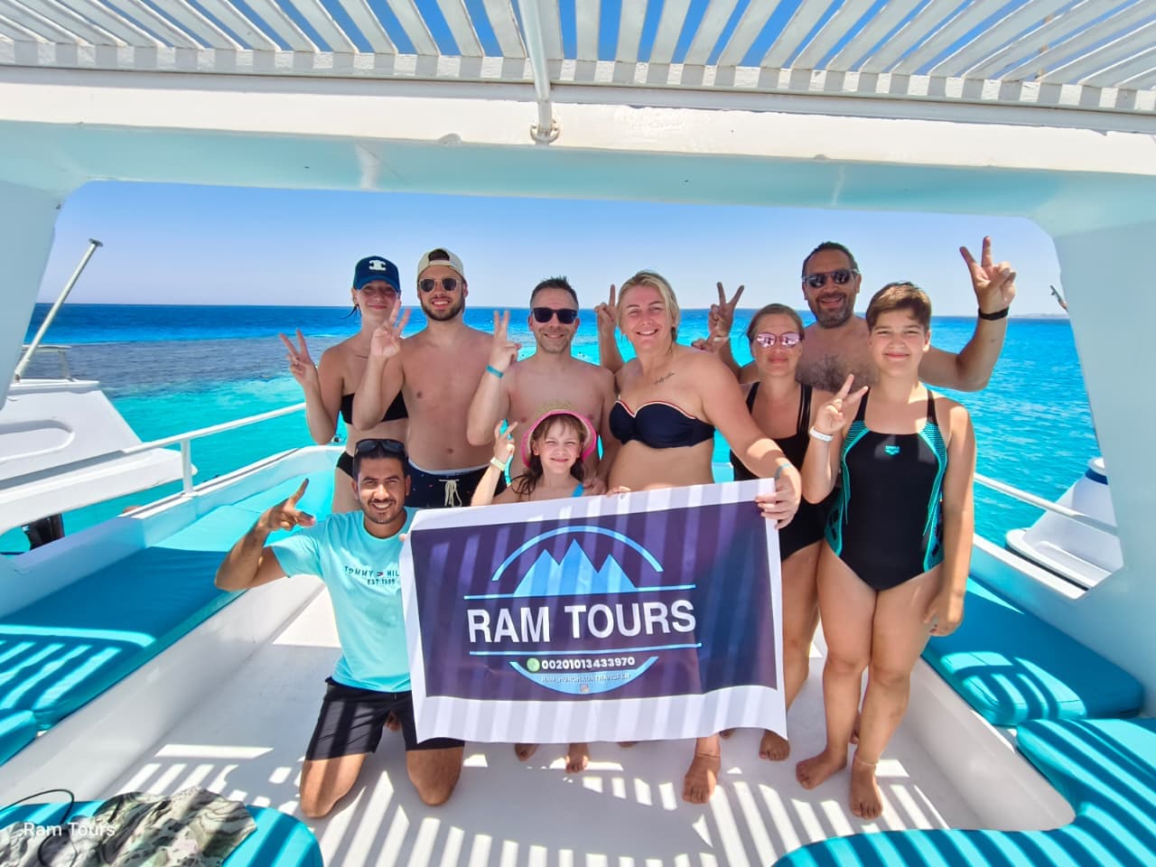 Smiling tourists holding a Ram Tours banner on a boat deck over clear blue water.